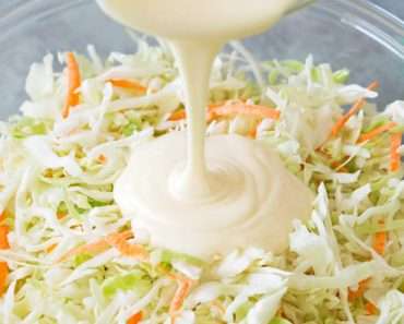 Um molho branco cremoso sendo despejado sobre uma salada de repolho dentro de uma travessa de vidro.