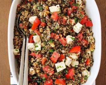 Uma salada de lentilha para o ano novo com quinoa, tomate e pedaços de queijo, servido dentro de uma travessa retangular branca, sob uma mesa de madeira.