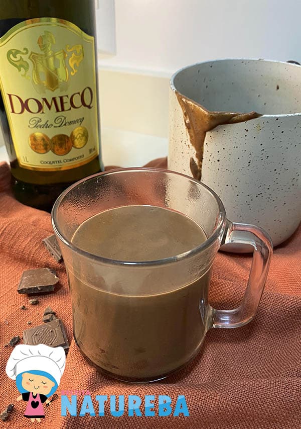 chocolate quente dentro de uma xícara sobre um pano vermelho, com uma garrafa de conhaque atras e uma leiteira com chocolate quente